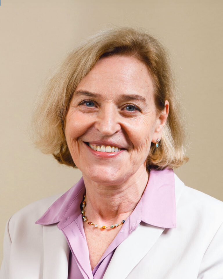 A smiling woman with shoulder-length blonde hair, wearing a white blazer over a light purple collared shirt and a beaded necklace.