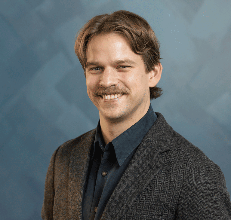 A man with a mustache smiling in a dark blazer and collared shirt.