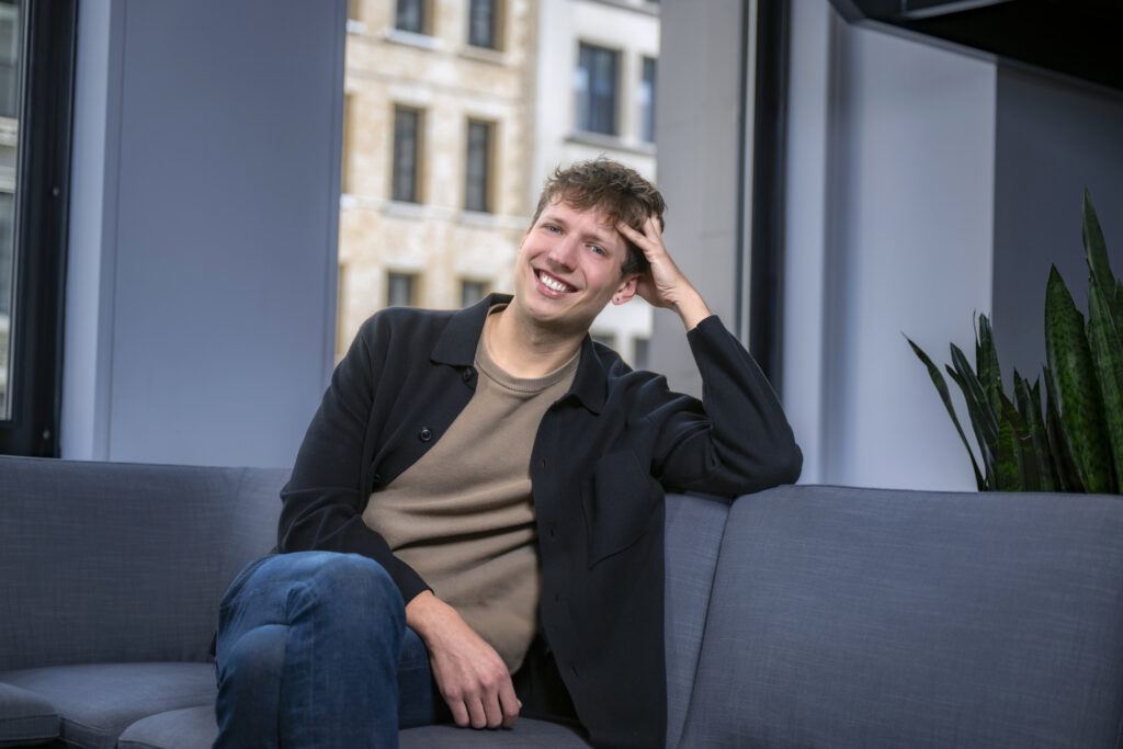 A smiling man sits on a grey sofa, one hand resting on his head.