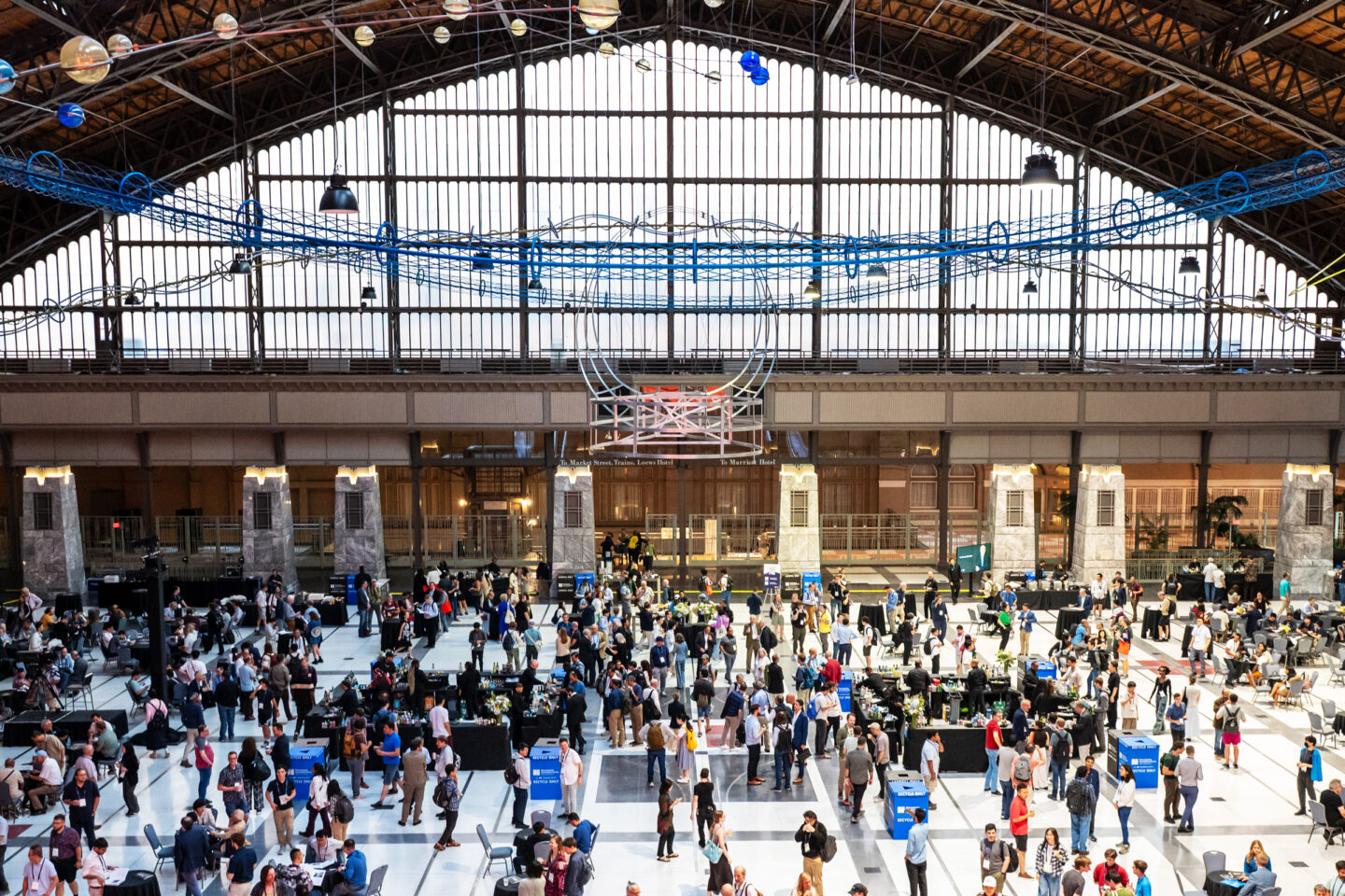 A large, airy transit hall filled with people attending an event at tables, beneath a high arched window and a large blue sculptural installation hanging from the ceiling. (credit: Emmages/Simons Foundation)