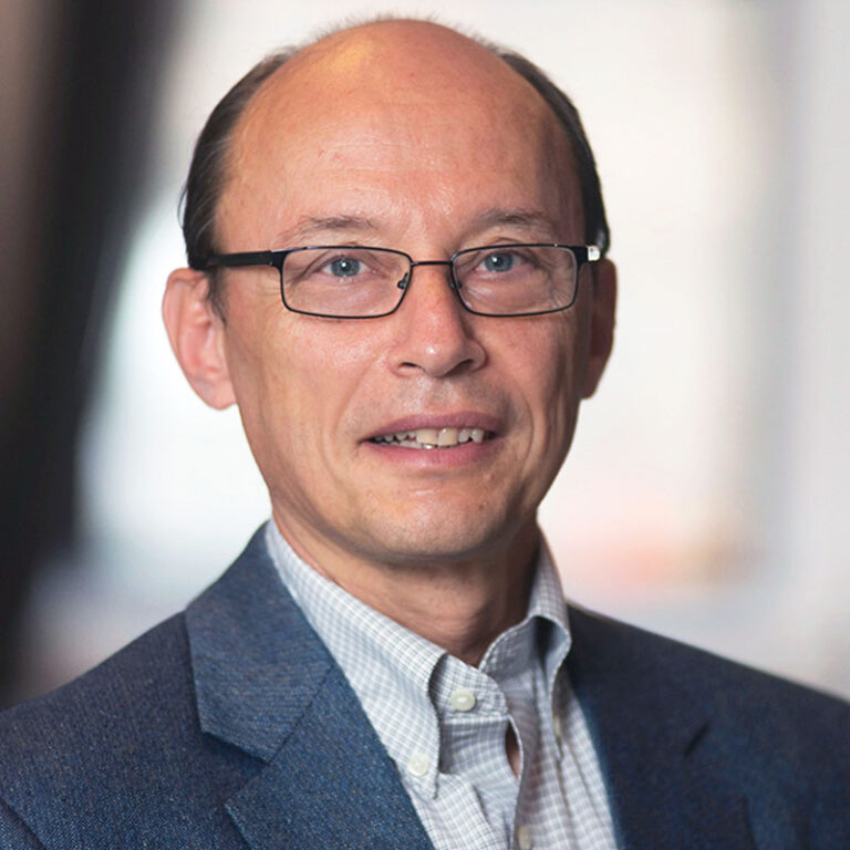A middle-aged man with glasses wears a blue blazer over a checkered button-down shirt.