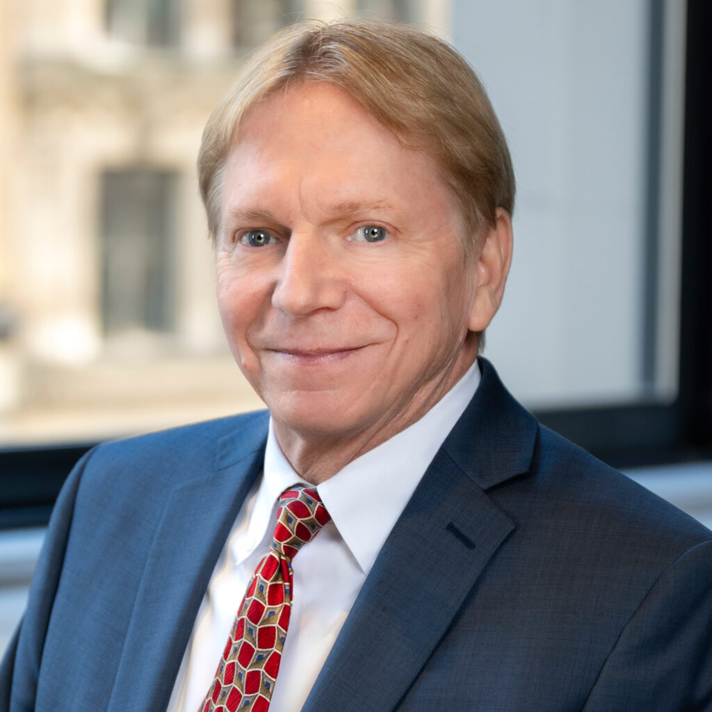 Professional headshot of a middle-aged man with blond hair wearing a blue suit, white shirt, and red patterned tie.
