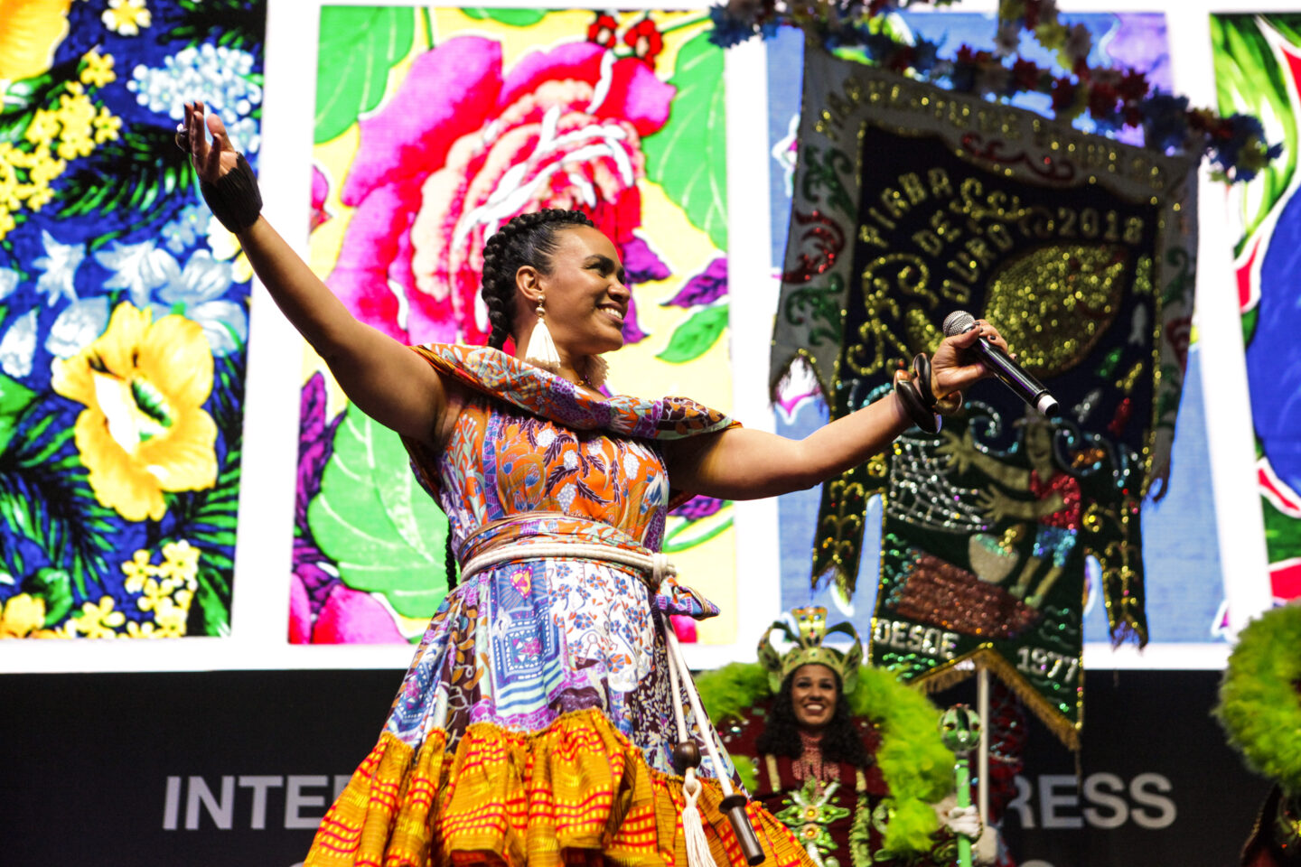 A woman in a vibrant patterned dress stands on stage with her arms raised, holding a microphone in front of large floral panels and a decorative banner.