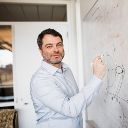 A man in a light blue button-down shirt writing on a whiteboard covered in mathematical equations.