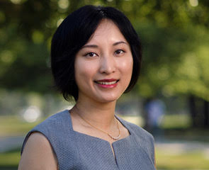 A portrait of a smiling woman with dark hair wearing a grey sleeveless top and a delicate gold necklace.