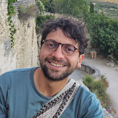 A man with dark curly hair, a beard, and glasses smiles at the camera in an outdoor setting.