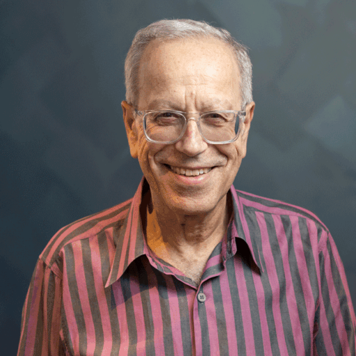 An older man with gray hair and glasses smiling at the camera, wearing a striped shirt.