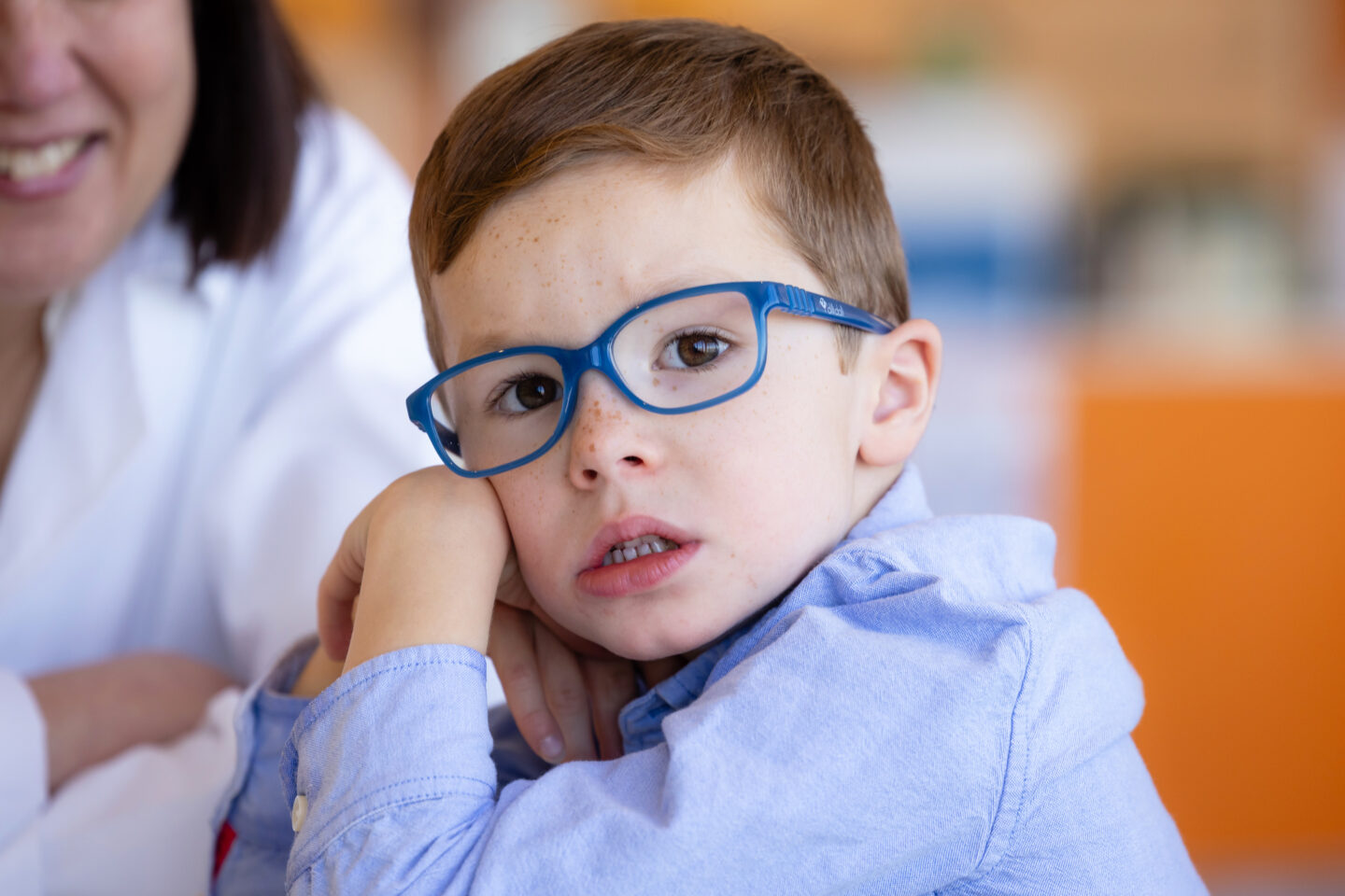 A young boy wearing blue glasses leans his face on his hand while sitting next to an adult.
