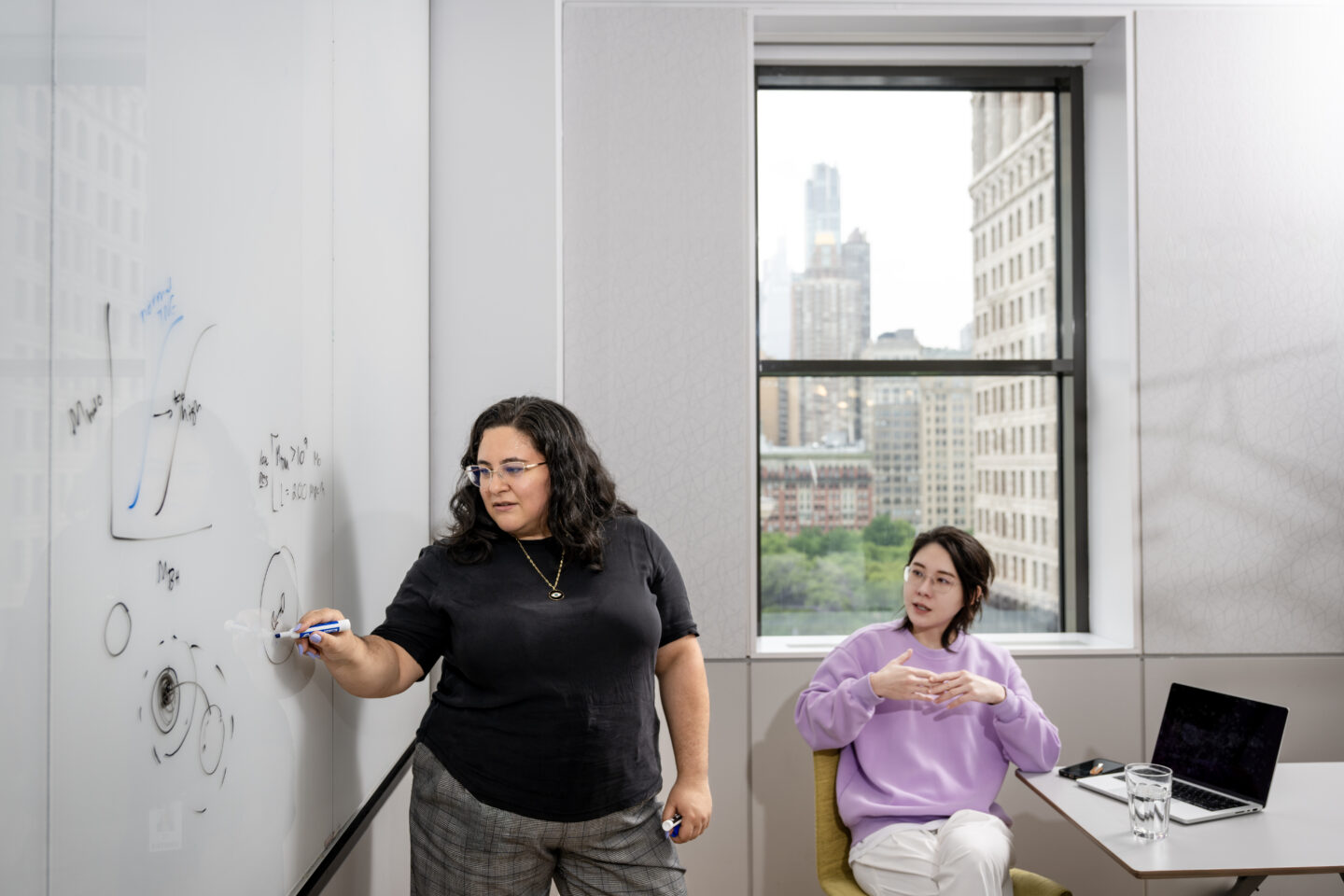 A woman writes on a whiteboard while another woman sits at a nearby table in an office with a city view.