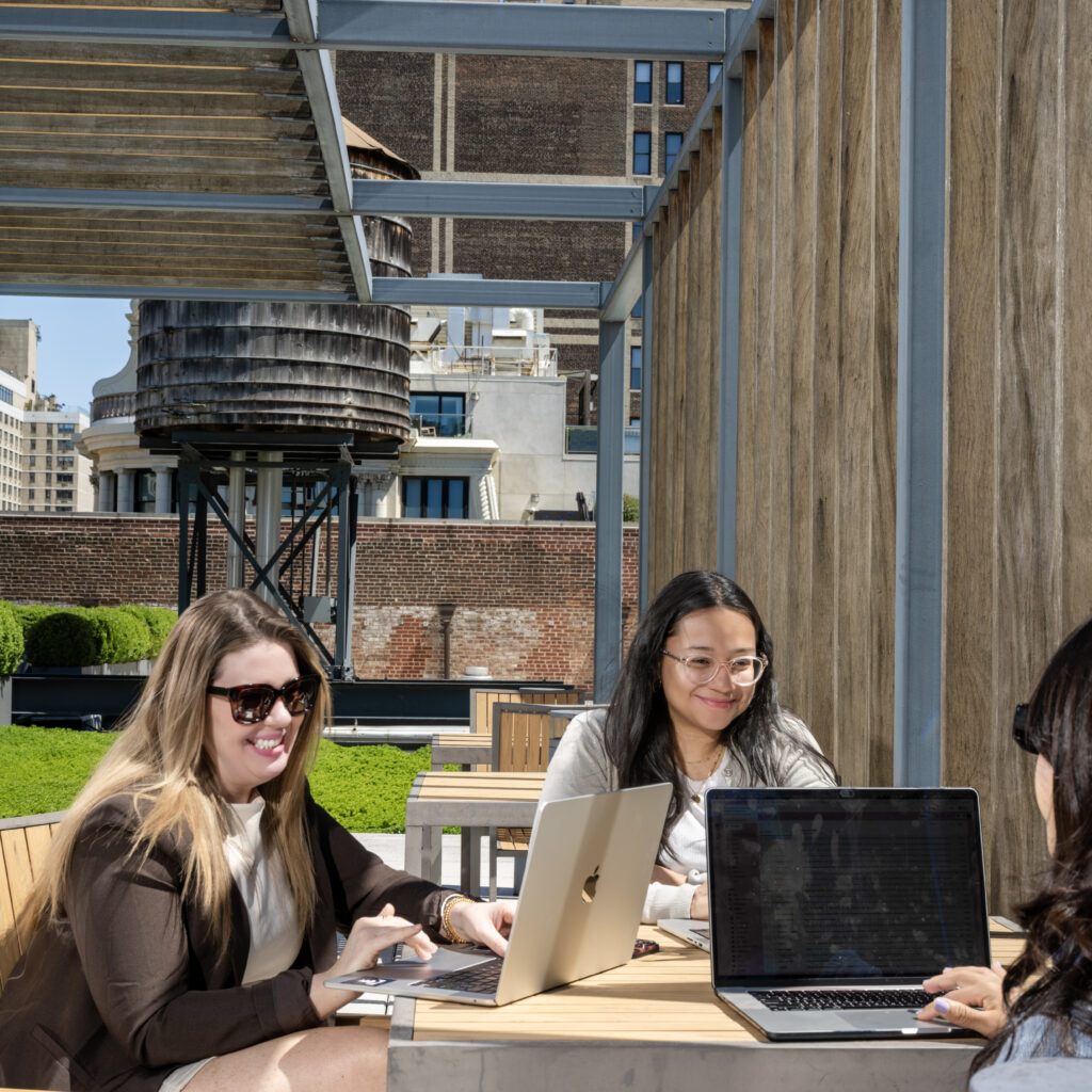 Three women work on laptops at an outdoor wooden table on a rooftop terrace.