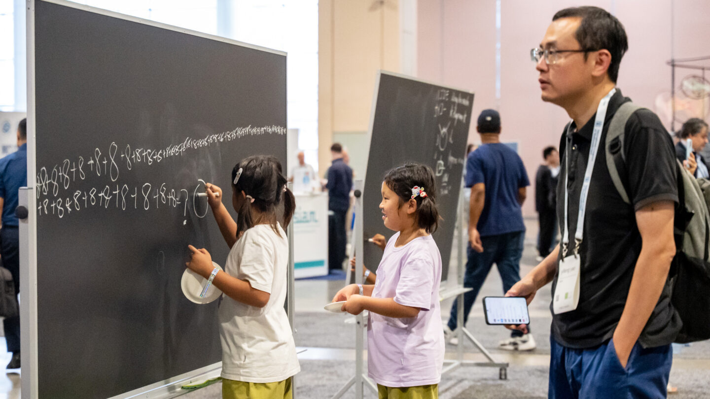 Two young girls write a long addition equation on a large chalkboard at a conference, while a man looks on.