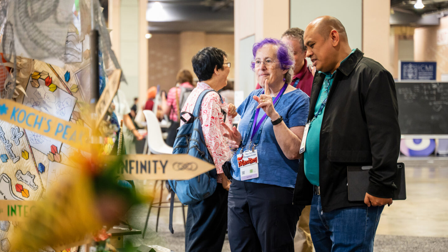 A woman with purple hair gestures while speaking to a man at a conference, with a decorative display featuring the text Kochs Peak and Infinity in the foreground.