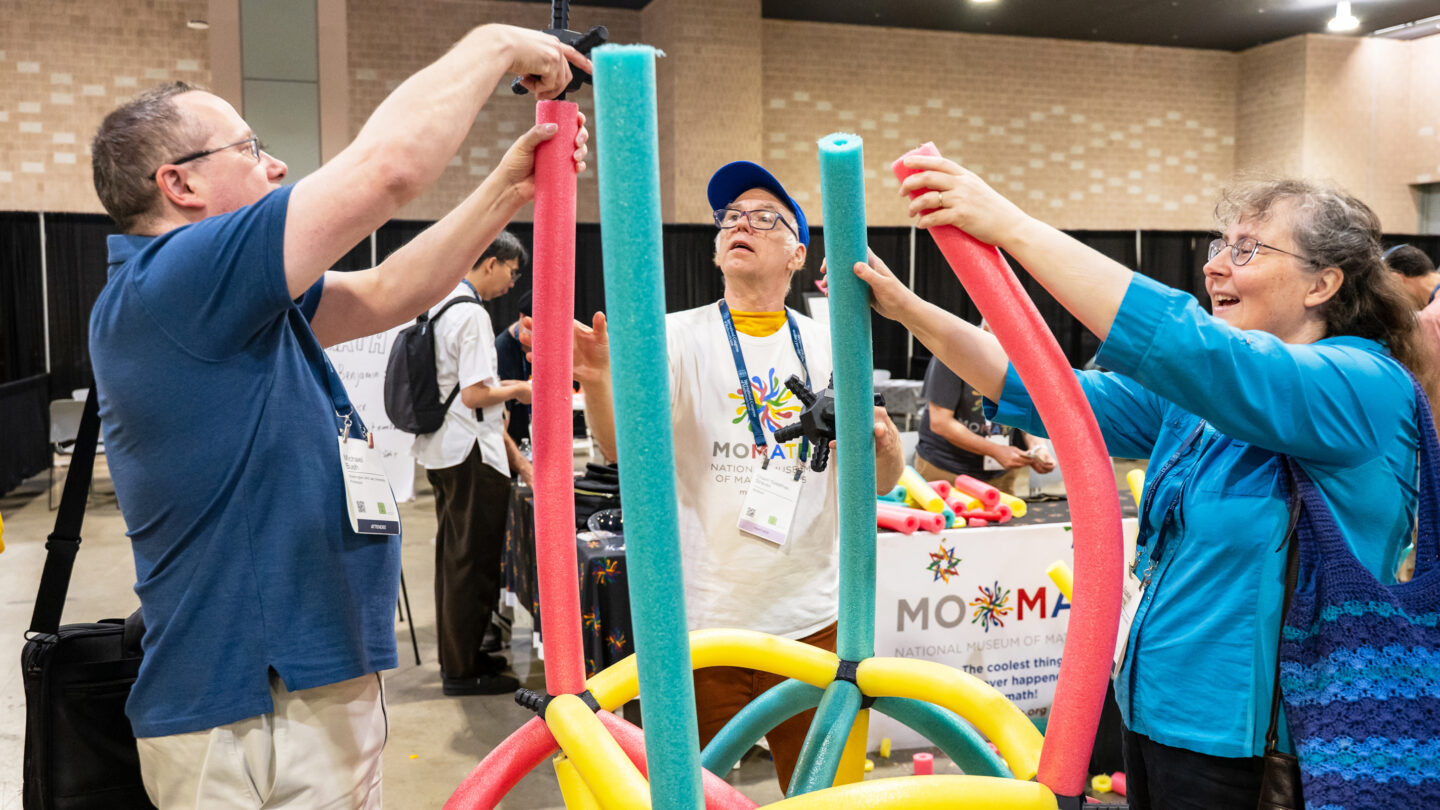 Two people build a structure using colorful foam pool noodles and plastic connectors at a National Museum of Mathematics event.