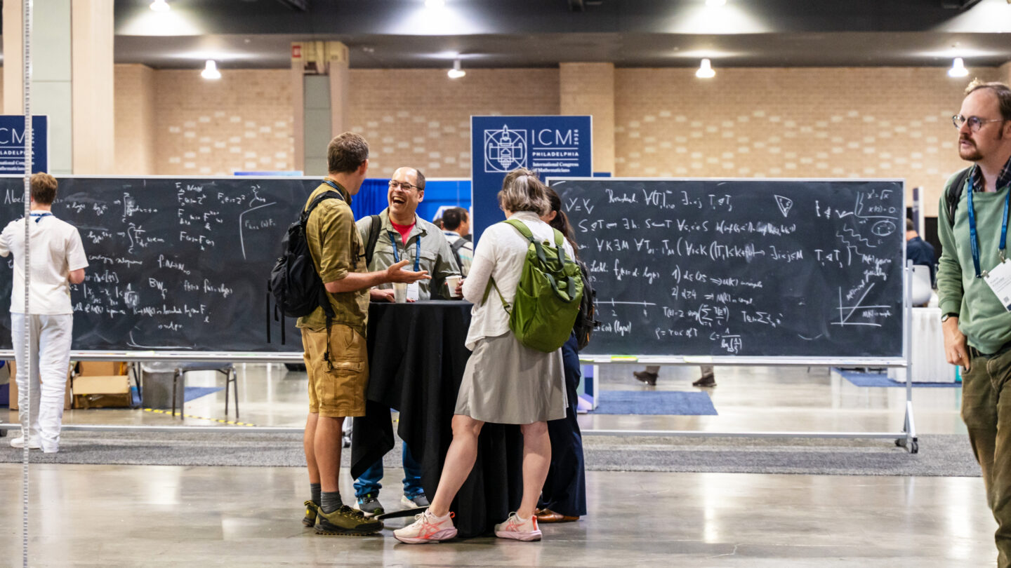 Three people stand around a high table at a conference, laughing and chatting, in front of large blackboards covered in mathematical equations.