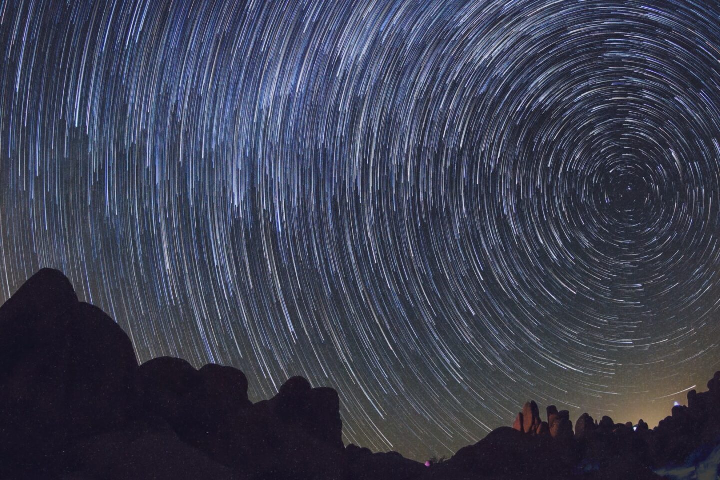 Long exposure star trails forming concentric circles in the night sky above a dark, jagged mountain silhouette.