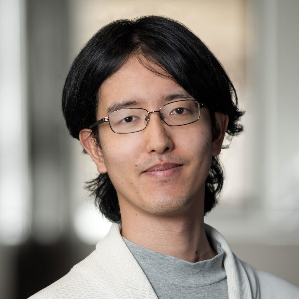 Portrait of a young man with dark hair and glasses wearing a light-colored cardigan.