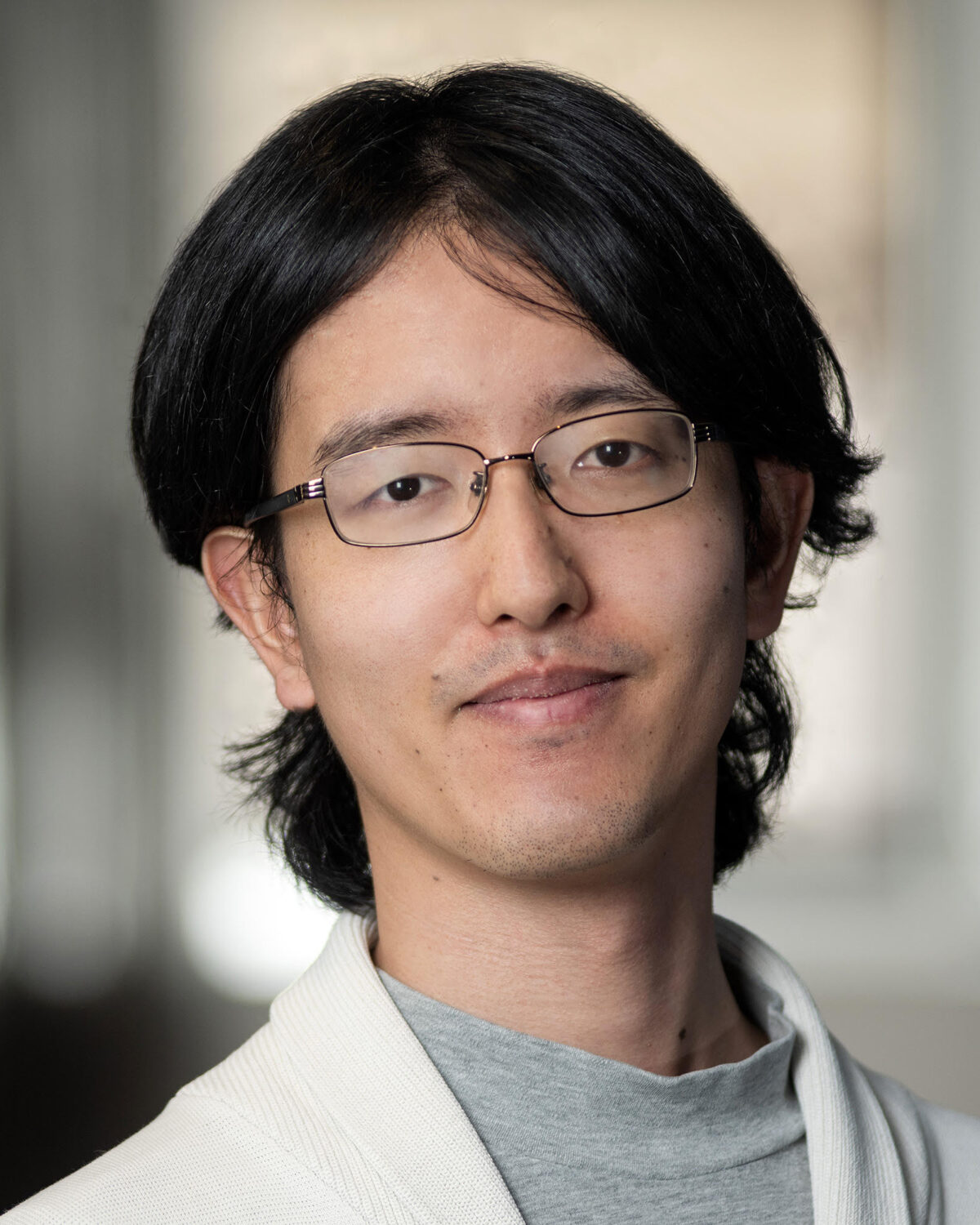 Portrait of a young man with dark hair and glasses wearing a light-colored cardigan.