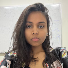 A portrait of a young woman with long, dark hair, wearing a patterned top in front of a whiteboard.