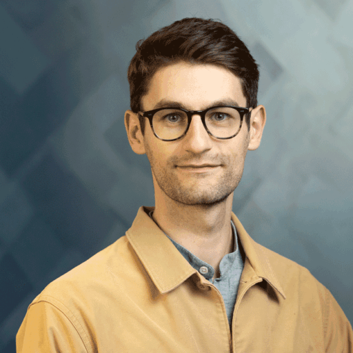 A man wearing glasses and a light-colored collared shirt against a patterned blue background.