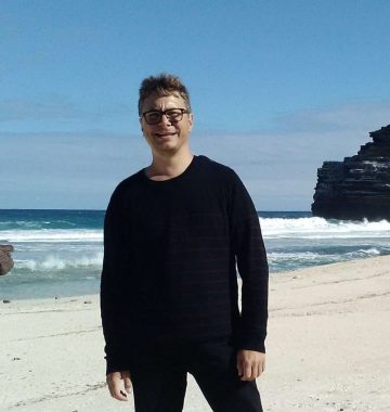 A man wearing glasses and a black long-sleeved shirt stands on a beach with the ocean and a rocky cliff in the background.