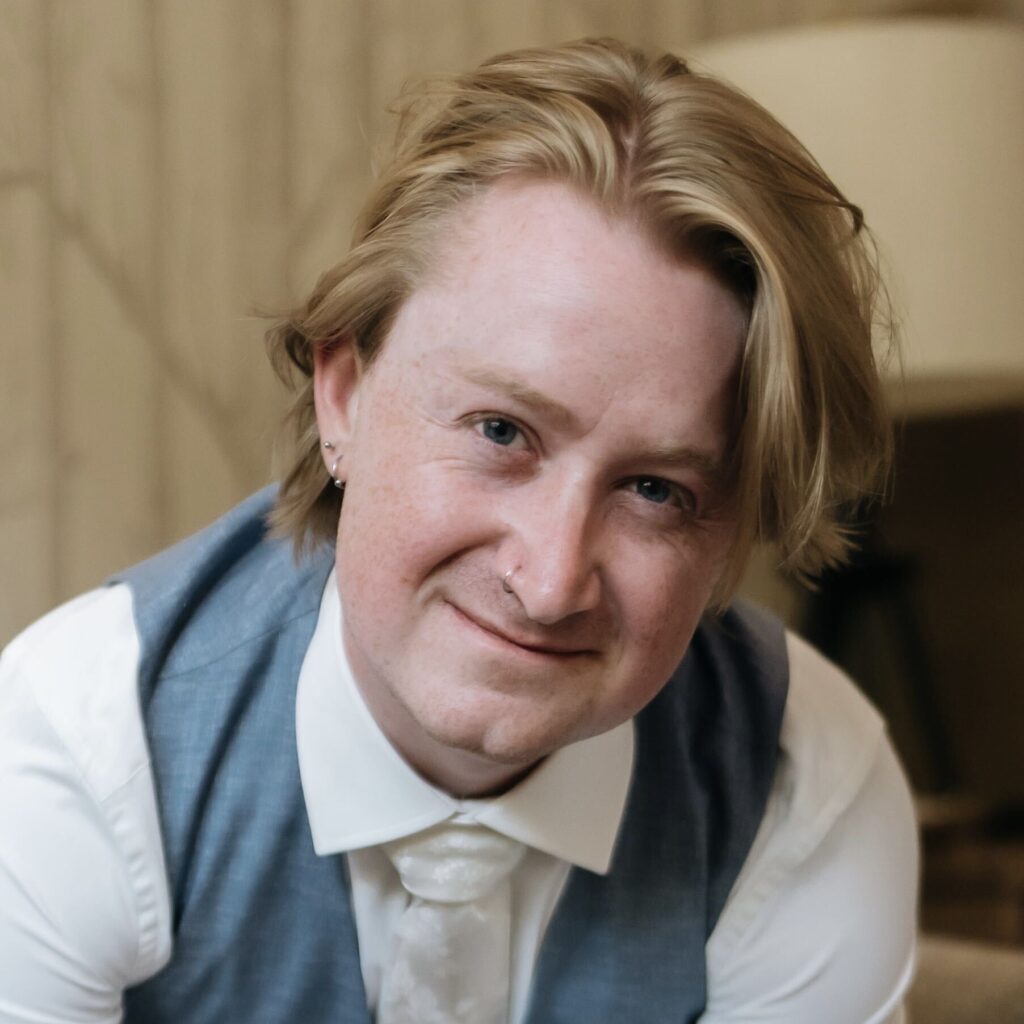 A portrait of a person with blonde hair wearing a white collared shirt, tie, and blue vest.