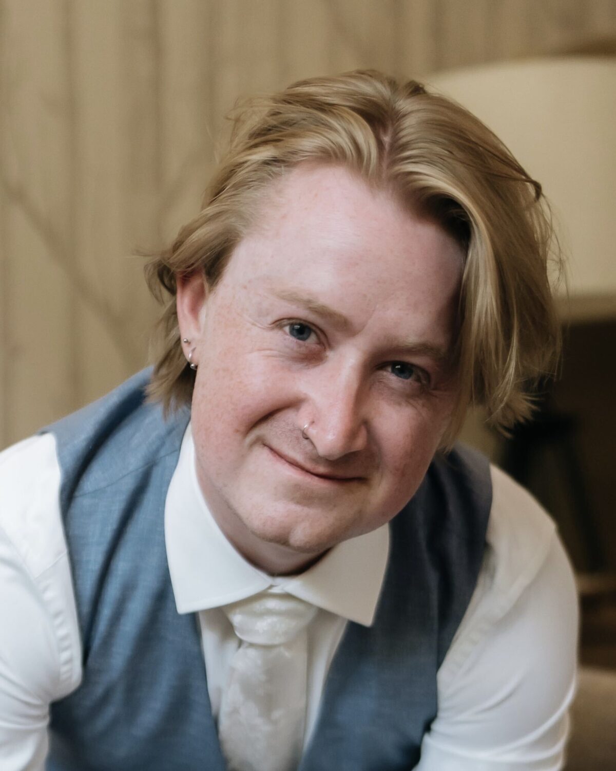 A portrait of a person with blonde hair wearing a white collared shirt, tie, and blue vest.